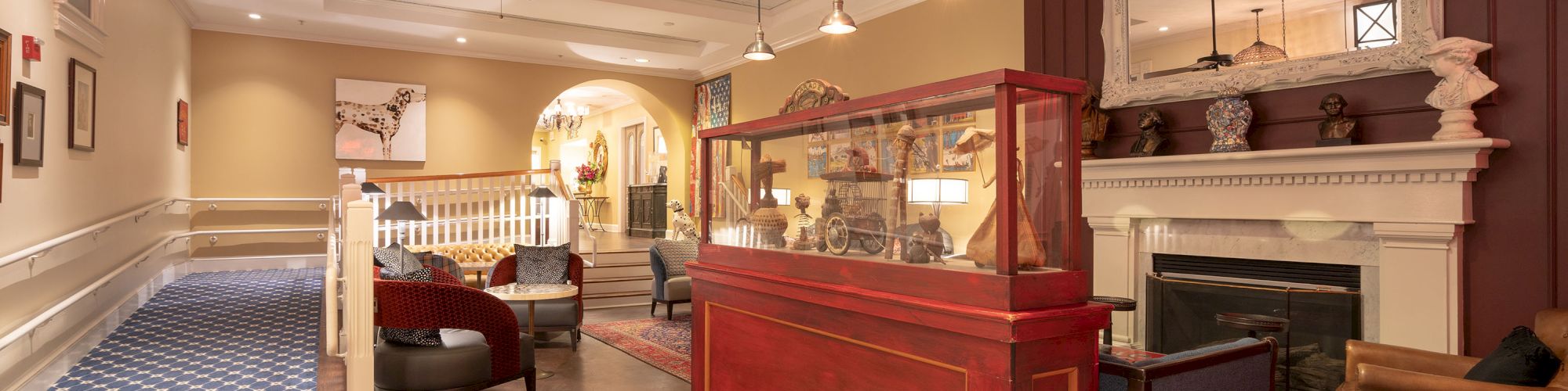 A warmly lit hotel lobby features a reception desk, cozy seating areas, a large mirror above a fireplace, and patterned carpets throughout the space.