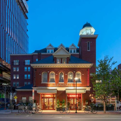 The image depicts a charming, well-lit brick building with a clock tower, located on a city street at dusk.