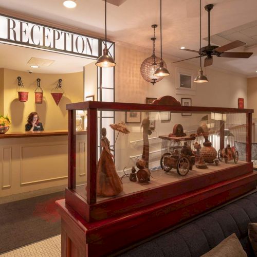 A reception area with a staff member behind the desk, decorative items in a glass display case, cozy seating, and a well-lit, welcoming atmosphere.