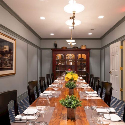 A dining room with a long wooden table, 12 chairs, and potted plants. There are place settings, hanging lights, a framed picture, and a cabinet in the back.